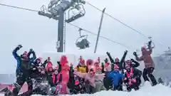 A large group of colorful skiers celebrate in deep snow beneath a foggy ski lift, arms raised in excitement on a snowy day.
